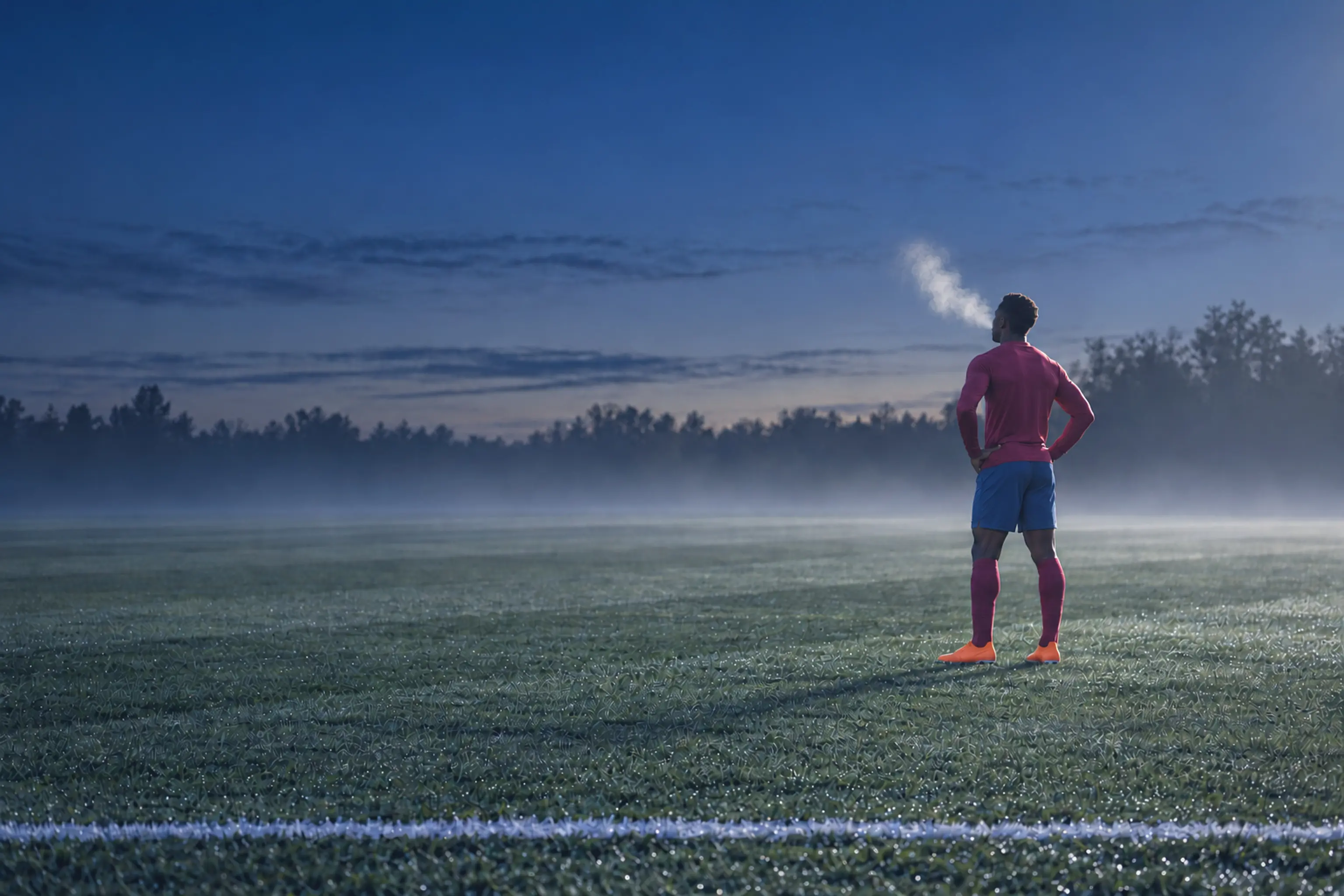 A footballer alone on a striped training pitch at first light, his breath lit by the floodlights
